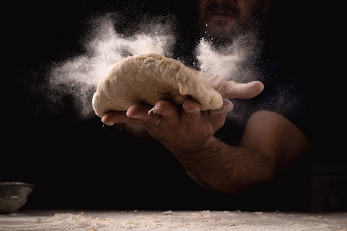Man throwing pizza dough in the air with flour clouds, symbolizing traditional food concepts abroad