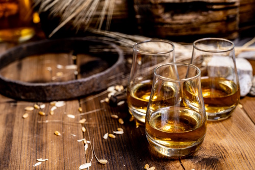 Three glasses of whisky on a rustic wooden table with barley and distilling elements in the background.